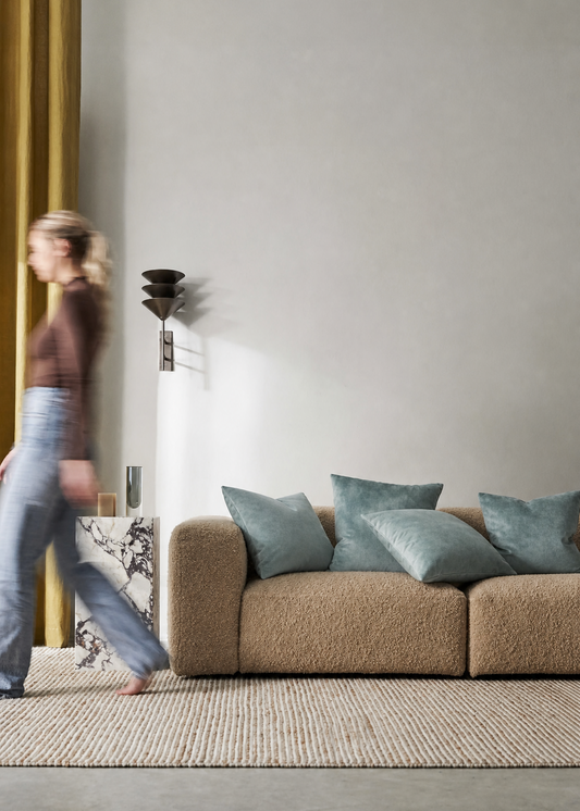 Woman walking past a beige sofa with decorative pillows in a modern living room.