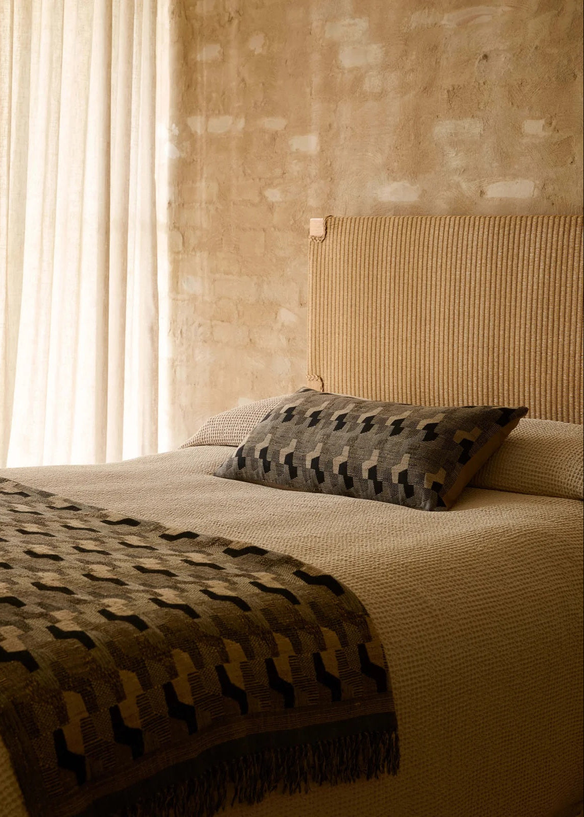 A styled bedroom scene featuring a bed with textured beige bedding, a geometric patterned throw blanket, and a matching accent pillow, set against a rustic plaster wall and sheer curtains.
