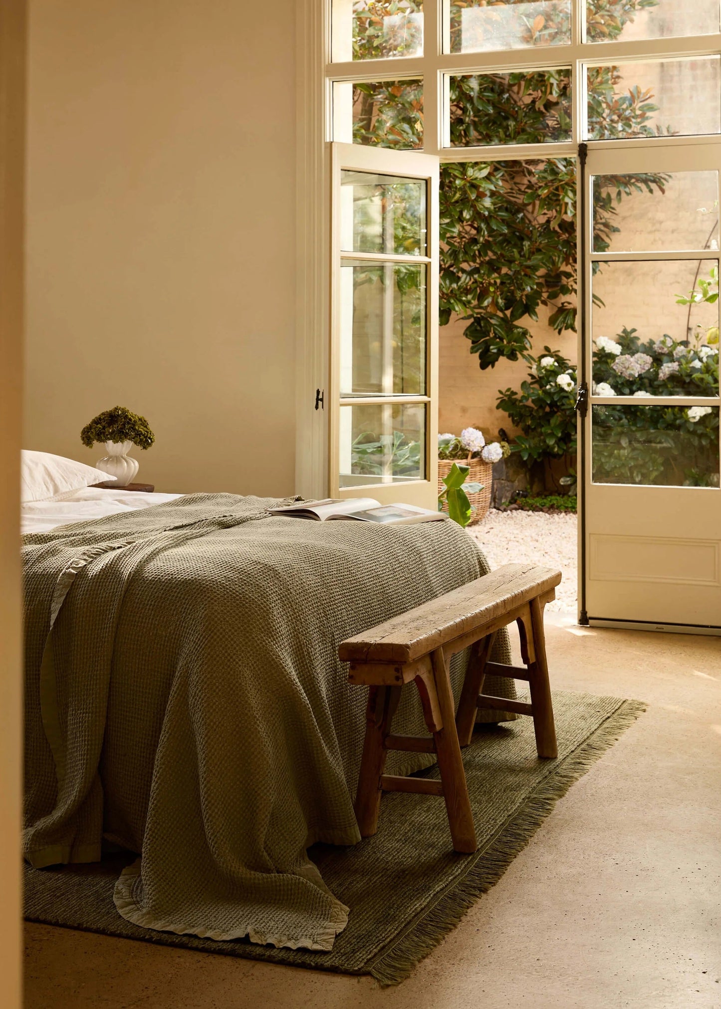 A cozy bedroom scene with a bed dressed in a textured olive-green blanket, a wooden bench at the foot of the bed, and large white-framed glass doors opening onto a sunlit garden courtyard.