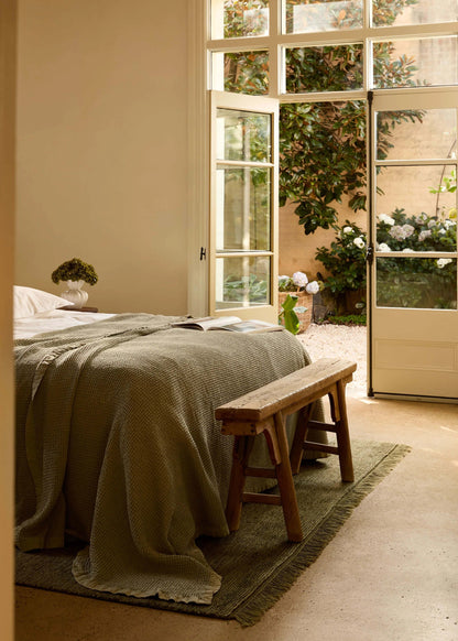 A cozy bedroom scene with a bed dressed in a textured olive-green blanket, a wooden bench at the foot of the bed, and large white-framed glass doors opening onto a sunlit garden courtyard.