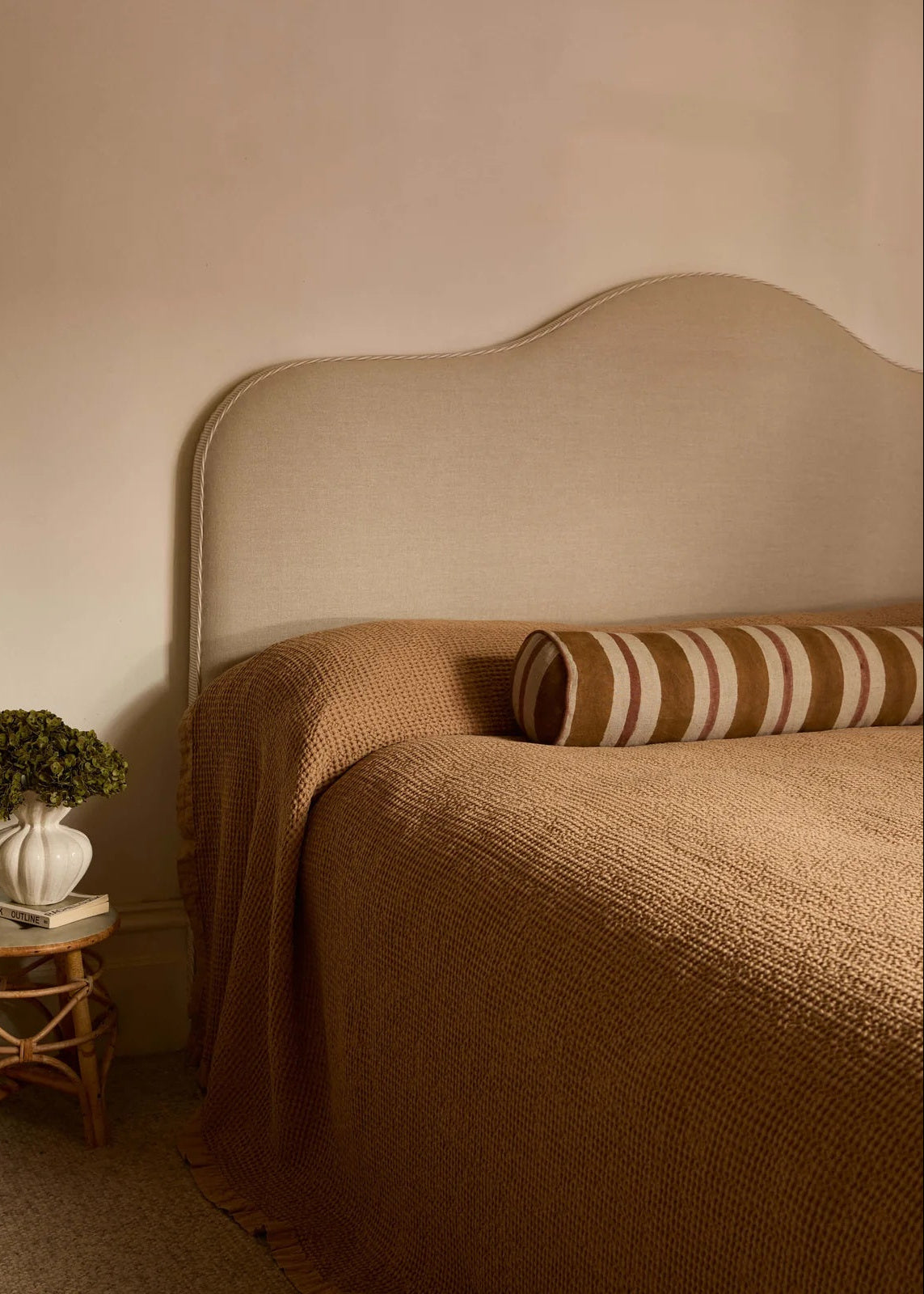 A cozy bedroom scene featuring a bed with a textured brown knit blanket, a matching scalloped upholstered headboard, and a striped brown and cream bolster pillow. A small wooden side table with a white vase of greenery sits to the left against a warm beige wall.