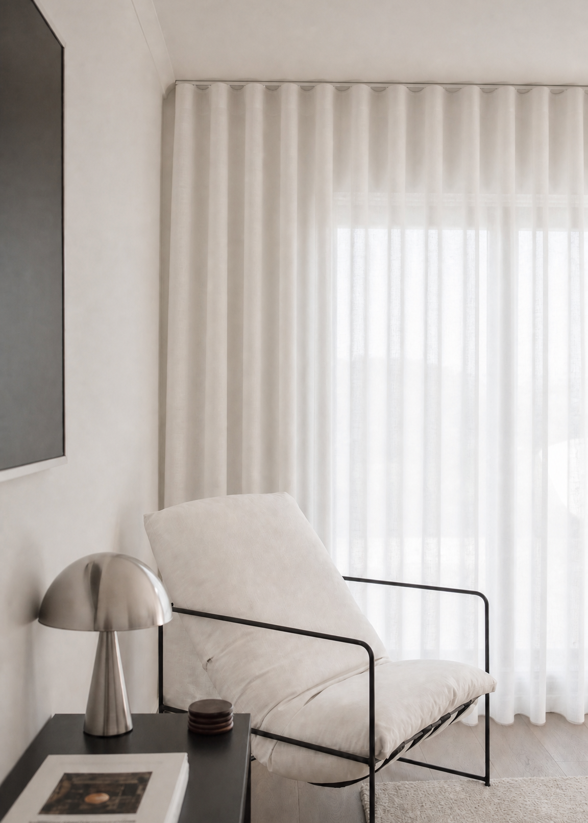 A modern white upholstered accent chair with a black metal frame, positioned next to a dark side table with a silver mushroom lamp and books, in front of a window with white sheer curtains.
