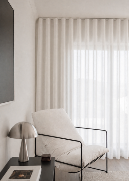 A modern white upholstered accent chair with a black metal frame, positioned next to a dark side table with a silver mushroom lamp and books, in front of a window with white sheer curtains.