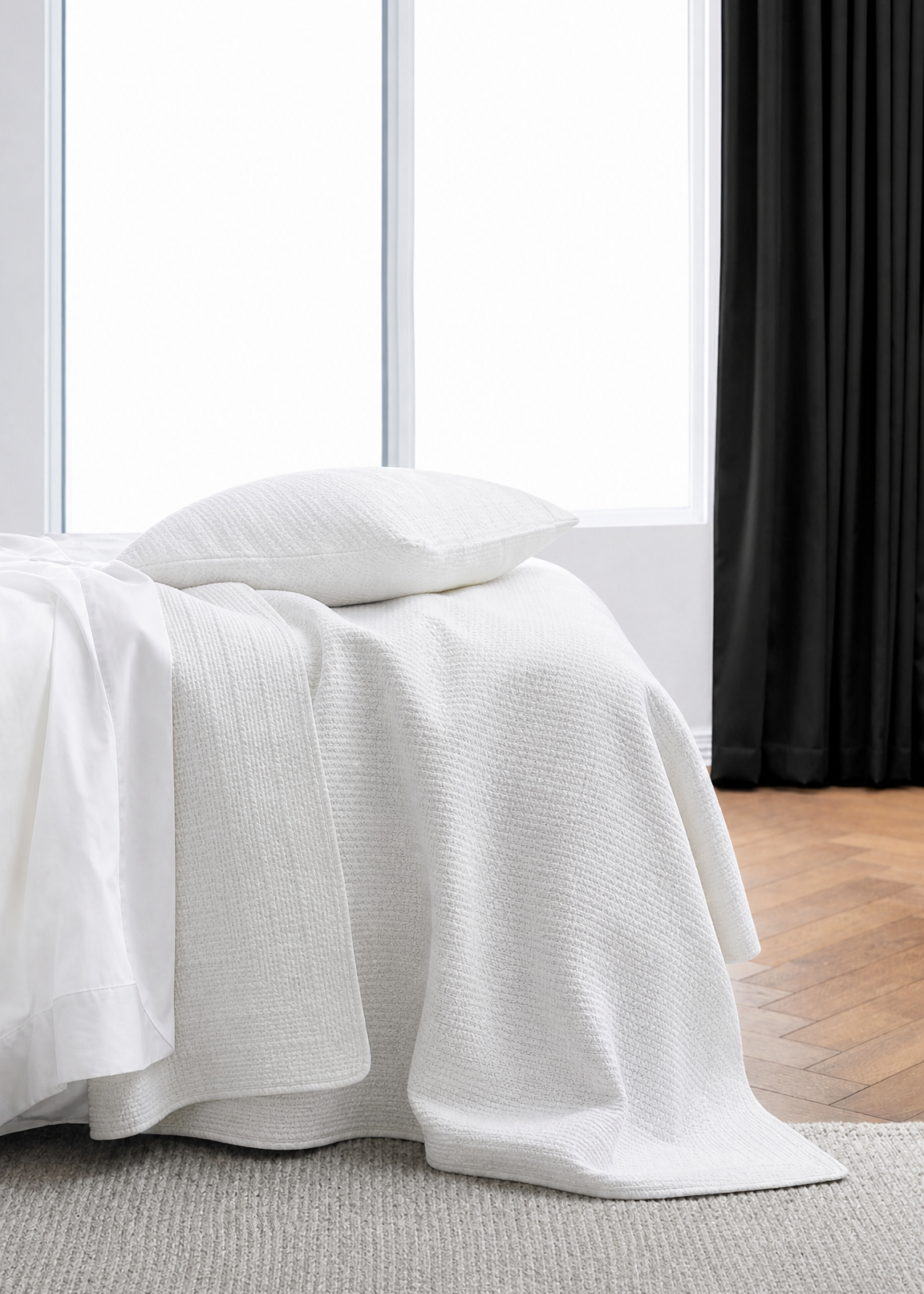 White textured blanket draped over a bed in a room with a window and black curtain.