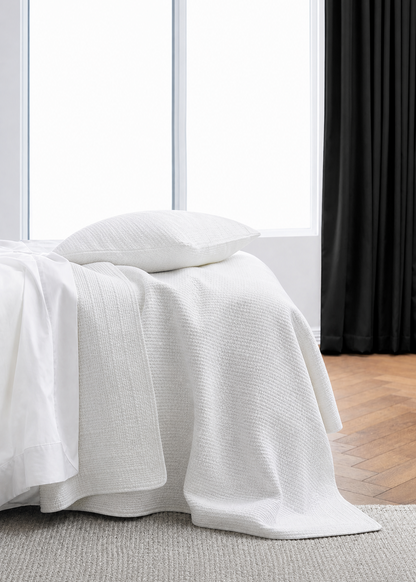 White textured blanket draped over a bed in a room with a window and black curtain.