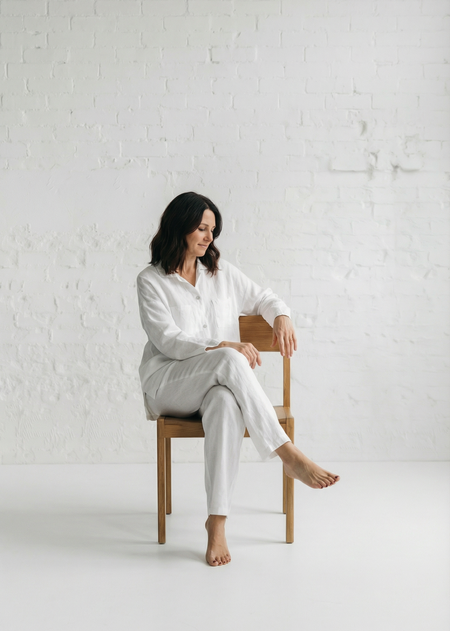 Woman in a white outfit sitting on a wooden chair against a white brick wall.