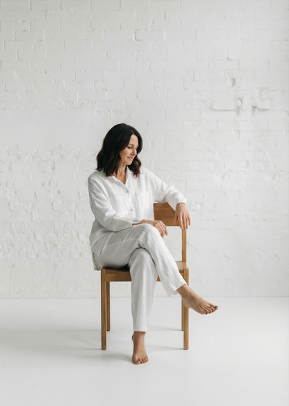 Woman in a white outfit sitting on a wooden chair against a white brick wall.
