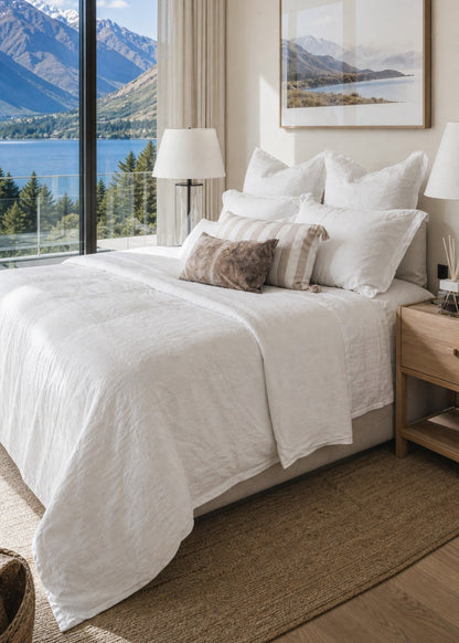 A neatly made bed with white bedding and decorative pillows in a bright bedroom, featuring a large window with a scenic mountain and lake view, a wooden nightstand, and a framed landscape print on the wall.