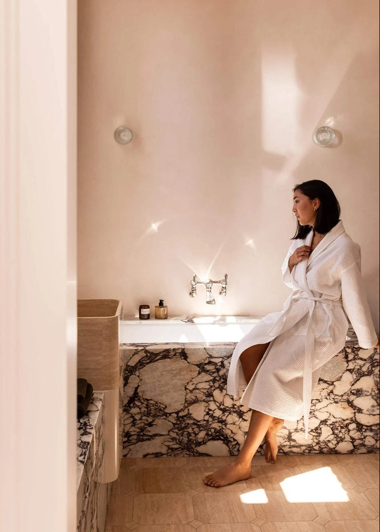 A woman in a white bathrobe sits on the edge of a marble bathtub in a warm, sunlit bathroom with beige walls and tiled floors.