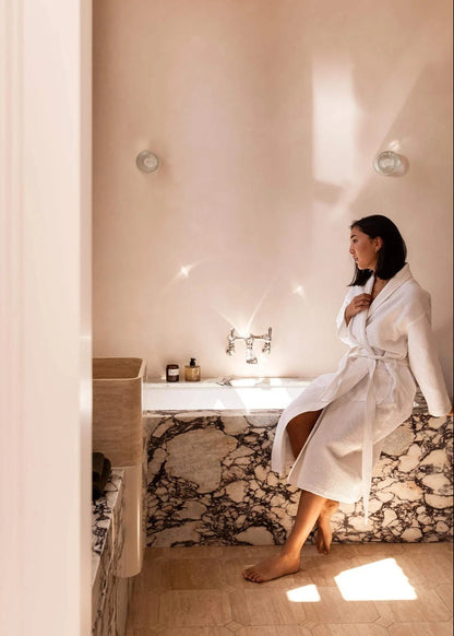 A woman in a white bathrobe sits on the edge of a marble bathtub in a warm, sunlit bathroom with beige walls and tiled floors.