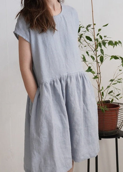 Woman wearing a light blue dress standing next to a plant on a stool against a plain background