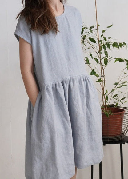 Woman wearing a light blue dress standing next to a plant on a stool against a plain background