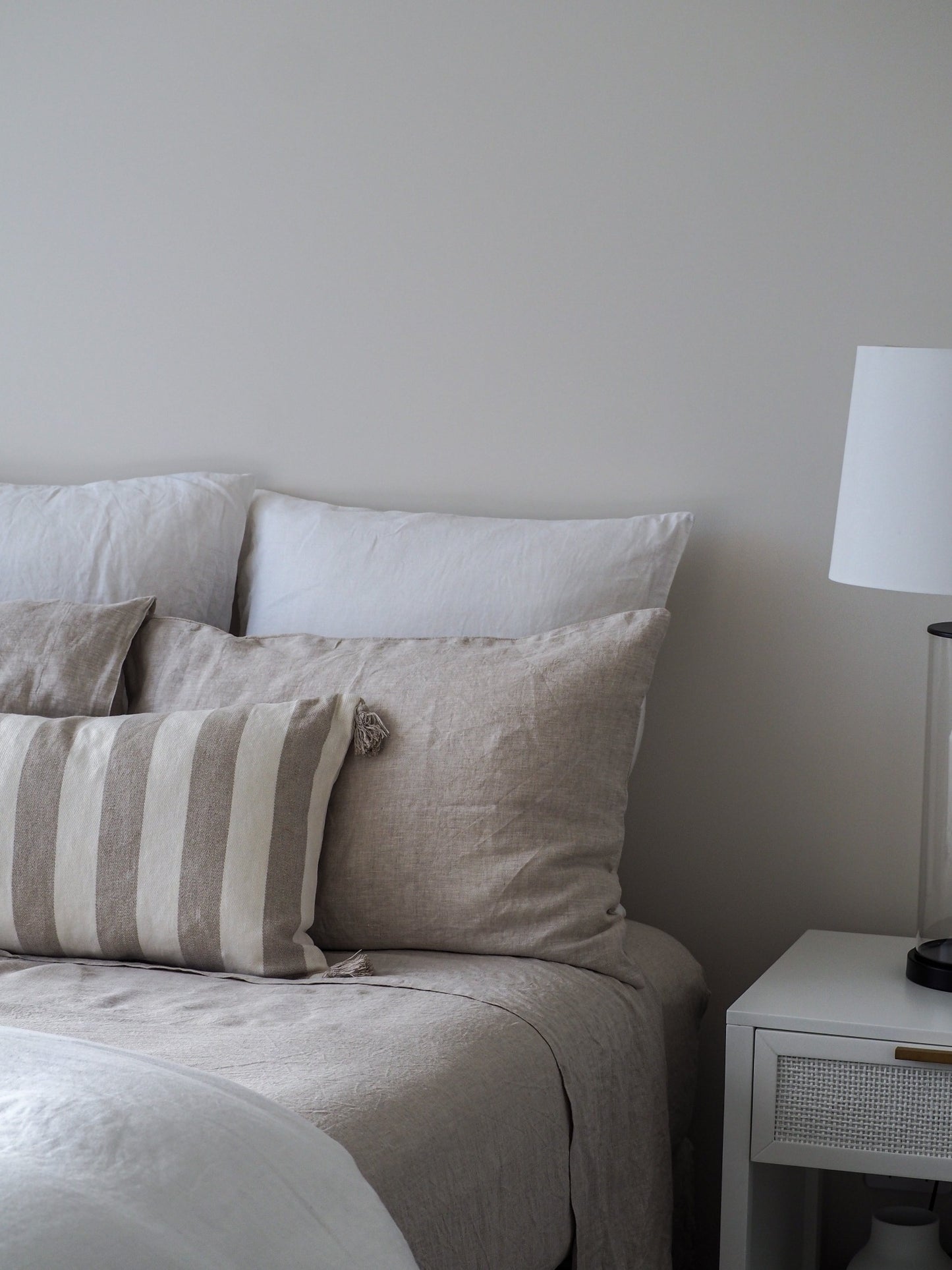 A neatly made bed with a beige linen sheet set, including a fitted sheet, a flat sheet, and pillowcases, alongside a nightstand with a lamp.