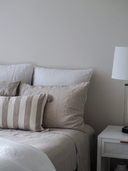 A neatly made bed with a beige linen sheet set, including a fitted sheet, a flat sheet, and pillowcases, alongside a nightstand with a lamp.